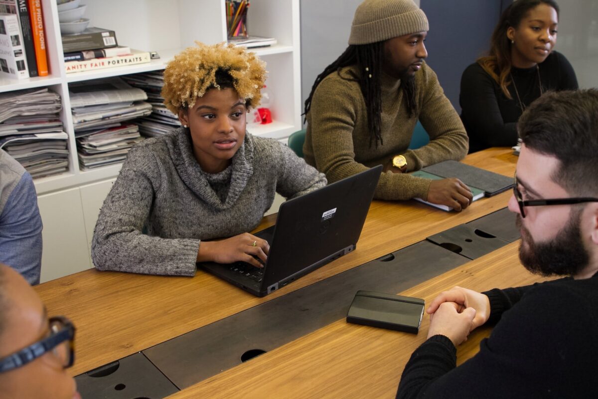 A group of individuals, men and women sitting at a large office desk with laptops